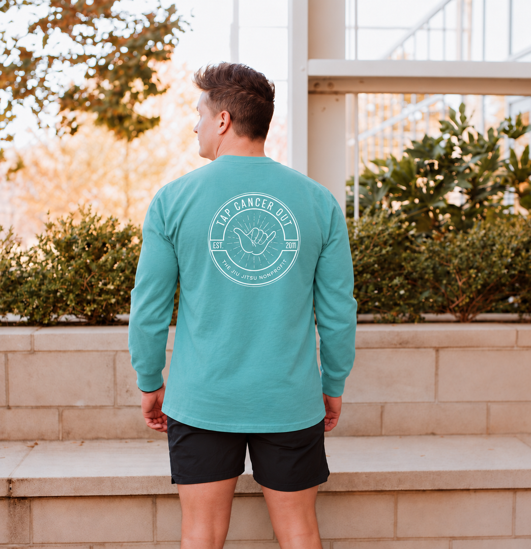 A man viewed from behind wearing a teal long-sleeve t-shirt with a white circular graphic on the back that reads 'Tap Cancer Out 
